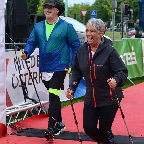 Ein Mann und eine Frau nehmen an einem Marathon teil, beide halten Skistöcke. Die Frau lächelt, während der Mann nach vorne schaut. Ein Banner hinter ihnen steht 'Nieder Osterr'.