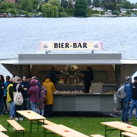 Eine Gruppe von Menschen versammelt sich um einen Food-Truck am Wasser, der mit 'Bier-Bar' beschriftet ist. Zwei Personen servieren Essen und Getränke im Inneren des Trucks, während andere warten. Holzbänke sind davor aufgestellt.
