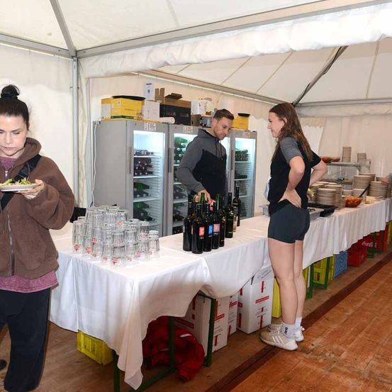 Ein Mann und zwei Frauen in einem Zelt. Der Mann hält Flaschen und einen Teller, die Frauen beobachten. Gläser, Kisten und Teller stehen auf den Tischen.