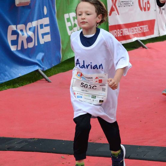 Ein junger Junge namens Adrian, der ein weißes T-Shirt und schwarze Hose trägt, läuft einen Marathon. Er hält ein Papier mit seiner Nummer, 503C. Dahinter befinden sich Banner mit Text.