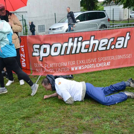 Ein Outdoor-Event mit Teilnehmern, einem großen roten Banner und einem Mann, der mit einem Stock vor ihm auf dem Boden liegt. Ein Mann mit einem Regenschirm steht hinter dem Banner.
