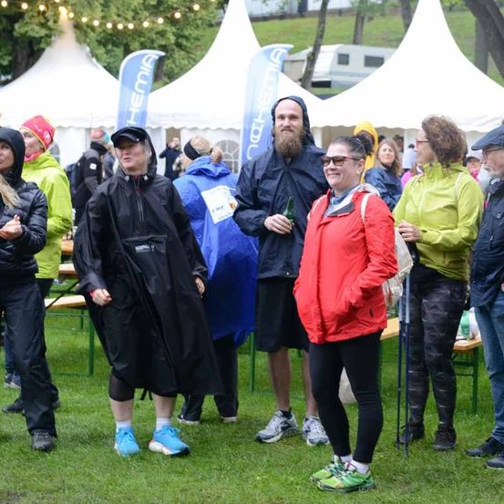 Eine Gruppe von Menschen in Regenmänteln und Turnschuhen steht auf einem Rasenfeld, möglicherweise bei einer Outdoor-Veranstaltung. Im Hintergrund sind einige Zelte zu sehen.