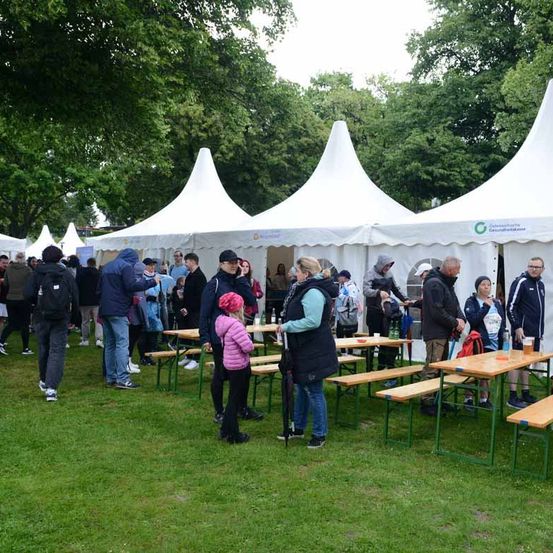 Viele Menschen versammeln sich unter weißen Zelten bei einer Veranstaltung im Freien. Einige sitzen an Bänken, andere stehen. Einige Personen tragen Hüte und einige von ihnen haben Getränke.