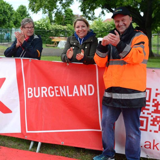 Drei Menschen stehen hinter einem roten Banner mit weißen Texten. Die Frau links klatscht. Der Mann in der Mitte lächelt und klatscht. Die Frau in der Mitte schaut etwas vor sich an.