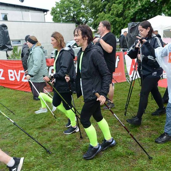 Eine Gruppe von Menschen in bunten Leggings und schwarzen Jacken geht mit Trekkingstöcken auf einem Rasenfeld. Dahinter steht ein Mann, der in ein Mikrofon spricht. Im Hintergrund befinden sich Zelte, ein roter Banner und ein Gebäude.