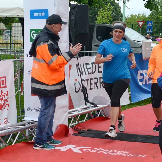 Ein Mann in einer orangefarbenen Weste spricht mit einer Frau, die auf einer Laufbahn läuft. Hinter ihnen befindet sich ein Banner mit der Aufschrift 'Niedere Osterreich'.