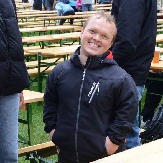 Ein Mann in einer schwarzen Jacke lächelt und steht in einem Sitzbereich im Freien mit Holzbänken und Tischen. Andere Menschen sitzen und stehen herum.