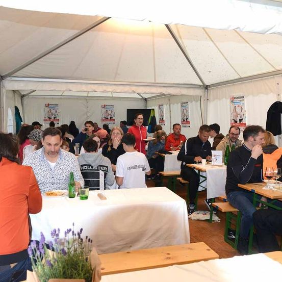 Eine Gruppe von Menschen sitzt in einem Zelt-Esssaal, einige tragen Brillen, Mützen und Sneakers. Eine Person steht vor dem Tisch und hält einen Teller. Der Tisch hat Teller, Weingläser und Flaschen. Das Zelt hat ein weißes Dach.