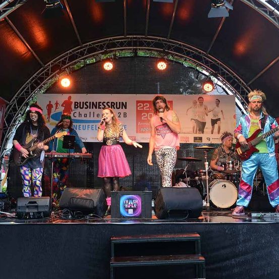 Eine Band tritt auf der Bühne auf, mit einem Banner im Hintergrund, das Businessrun Challenge 2B besagt. Zwei Frauen singen, während ein Mann ein Mikrofon hält. Es gibt Lautsprecher und Musikinstrumente auf der Bühne.
