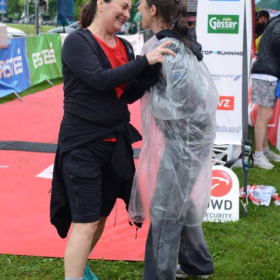 Zwei Frauen umarmen sich bei einer Veranstaltung im Freien. Die Frau auf der linken Seite trägt eine schwarze Jacke und ein rotes Shirt, während die Frau auf der rechten Seite in einem Regenponcho steht. Sie stehen auf einem roten Teppich.