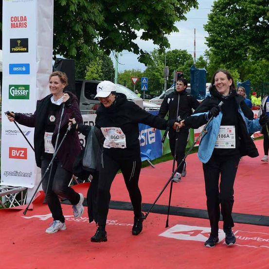 Drei Frauen beenden einen Marathon mit Skistöcken, lächelnd und sich an den Händen haltend. Sie tragen nummerierte Trikots. Hinter ihnen laufen zwei weitere. Banner, Bäume und geparkte Fahrzeuge sind im Hintergrund.