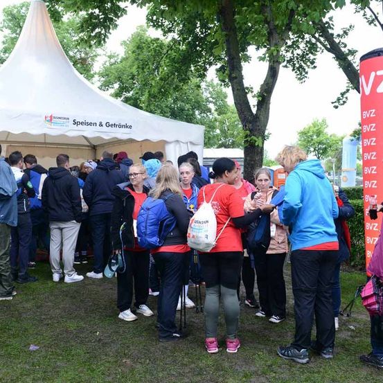 Eine Gruppe von Menschen steht in einer Schlange unter einem Zelt, möglicherweise bei einer Veranstaltung. Einige tragen rote T-Shirts und haben Rucksäcke dabei. Ein hoher roter Banner steht rechts.