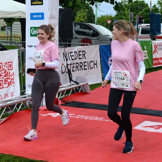 Zwei Frauen in rosa Shirts und Laufschuhen laufen auf einer roten Laufbahn mit Bannern im Hintergrund. Eine hält ein Telefon und eine Medaille, während die andere lächelt. Ein weißes Zelt und Autos sind im Hintergrund.