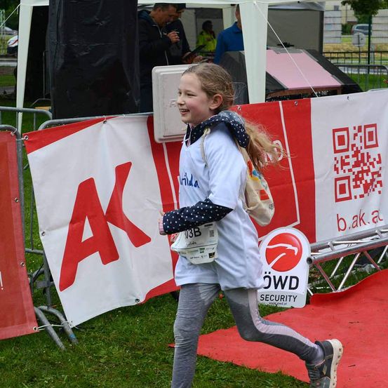 Ein junges Mädchen läuft mit fröhlicher Miene über ein Grasfeld und trägt ein Startnummernband. In der Nähe befindet sich ein roter Banner mit weißen Buchstaben und einem QR-Code.