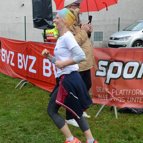Eine Frau in einem weißen Shirt läuft auf einem Rasenfeld mit einem roten Banner. Hinter ihr hält ein Mann einen Regenschirm und lächelt. Ein Auto ist in der Nähe geparkt.