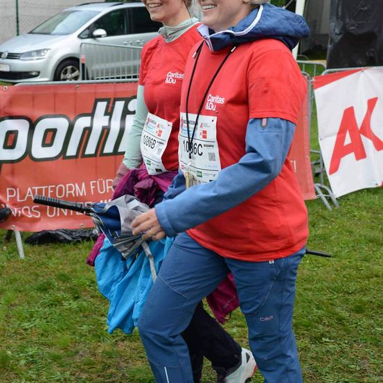 Zwei Frauen gehen in der Nähe eines Grasbereichs mit einem Banner hinter ihnen. Sie halten einen Regenschirm und tragen rote und blaue Shirts. Sie haben Nummernschilder an ihren Shirts. Ein Auto ist hinter ihnen geparkt.