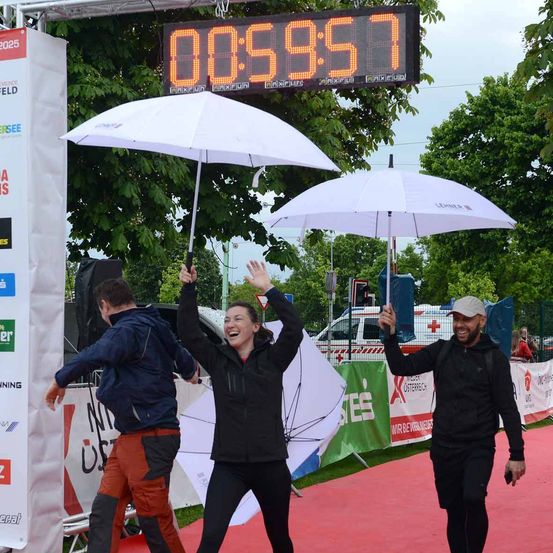 Drei Personen gehen auf einem roten Teppich und halten Regenschirme. Eine digitale Uhr zeigt die Zeit 00:59:57. Sie scheinen zu feiern. Hinter ihnen ist ein weißes Banner mit einem Logo. Bäume und ein Fahrzeug sind im Hintergrund.