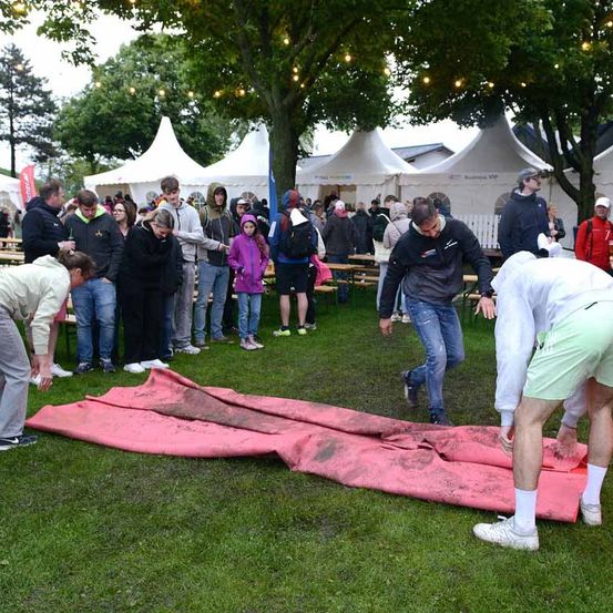 Mehrere Menschen versammeln sich in einem Park, einige sind gebeugt und untersuchen eine große rote Plane auf dem Boden. In der Nähe versammelt sich eine Menschenmenge in der Nähe weißer Zelte.