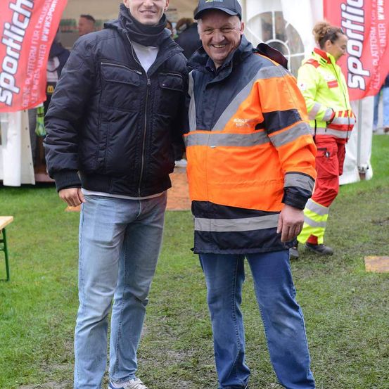 Zwei Männer stehen auf einem Rasenfeld und lächeln für ein Foto. Der Mann auf der linken Seite trägt eine schwarze Jacke und Jeans. Der Mann auf der rechten Seite trägt eine orange Sicherheitsjacke. Hinter ihnen steht eine Frau in einer gelben Sicherheitsjacke in der Nähe eines Zeltes.