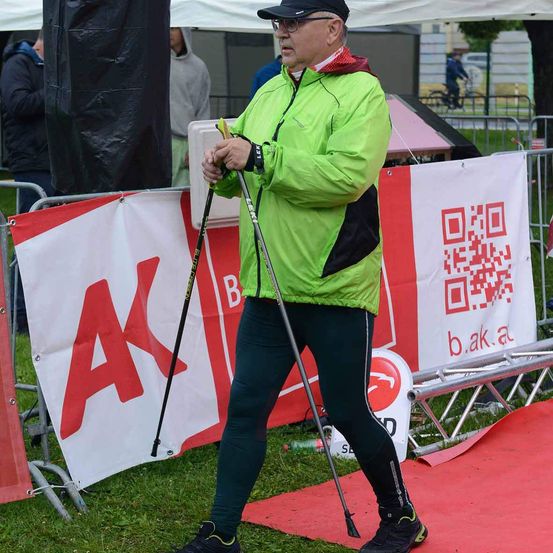 Ein Mann in einer grünen Jacke und schwarzen Hosen geht auf einem roten Teppich und hält Skistöcke. Hinter ihm befinden sich Banner mit den Buchstaben AK und B. Rechts ist eine Metallbarriere mit einem QR-Code zu sehen. Im Hintergrund sind Menschen und Fahrräder zu sehen.