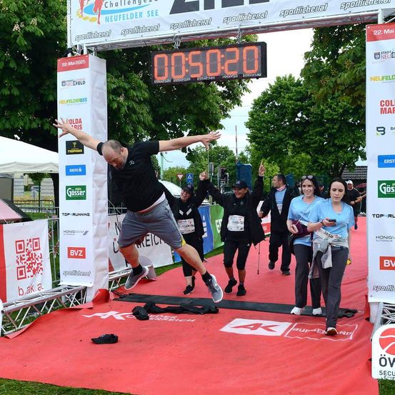 Ein Läufer in einem schwarzen Shirt und Shorts beendet einen Marathon und springt über eine Ziellinie, auf der ein digitales Timer 00:50:20 anzeigt. Hinter ihm laufen mehrere Leute, und eine Frau geht auf die Kamera zu. Im Hintergrund befinden sich Banner und Zelte.
