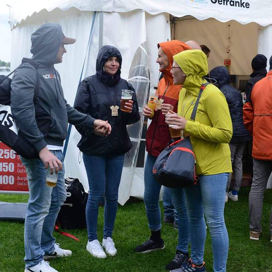 Eine Gruppe von Menschen steht in einem Außenbereich, wahrscheinlich auf einem Festival. Ein Mann schüttelt einer Frau die Hand, und eine Frau mit einer Tasche lächelt. Eine andere Frau hält ein Glas mit einem Getränk, und ein Mann hält ein Glas mit einem Getränk. Dahinter steht ein Zelt mit einem Schild, auf dem Gerranke steht.