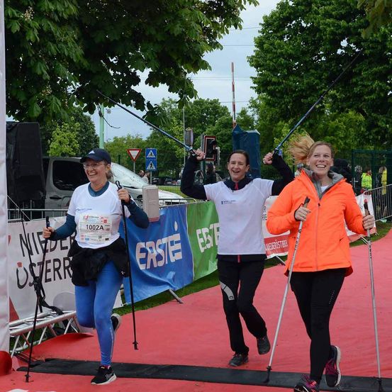 Drei Frauen laufen auf einer roten Laufbahn und halten Skistöcke in der Hand. Sie lächeln und scheinen den Wettkampf zu genießen. Die Laufbahn ist von Gras, Bäumen und einem Banner umgeben. Dahinter stehen ein Lieferwagen, ein Lautsprecher und ein Verkehrsschild.