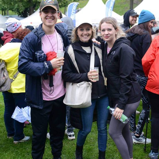 Eine Gruppe von Menschen steht auf einem Rasen. Ein Mann mit Hut hält einen Becher. Zwei Frauen stehen neben ihm, eine hält einen Becher und die andere eine Flasche. Sie alle lächeln. Dahinter sind Zelte und Menschen in Jacken.