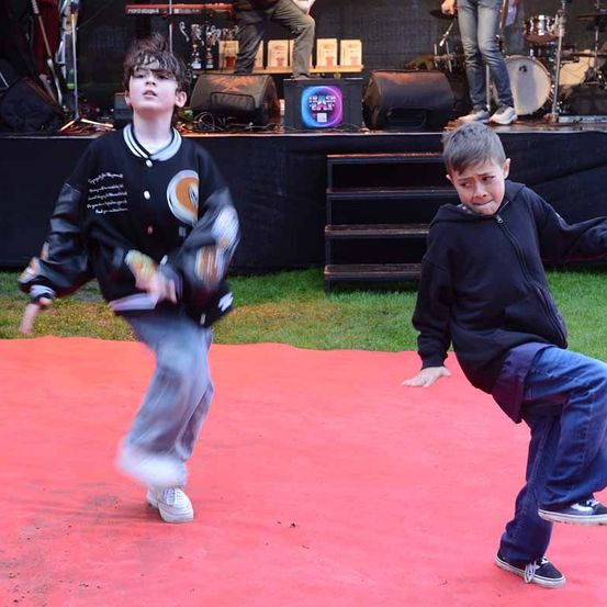 Zwei Jungen treten auf der Bühne auf, einer sieht verängstigt aus, während der andere begeistert tanzt. Dahinter steht eine Bühne mit Musikinstrumenten und Lautsprechern.