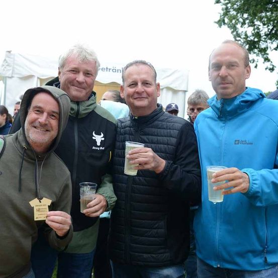 Vier Männer stehen zusammen und halten Getränke bei einer Veranstaltung im Freien, einer trägt eine grüne Kapuzenjacke, einer ein schwarzes Jackett und zwei blaue Jacken. Sie lächeln und posieren für ein Foto.