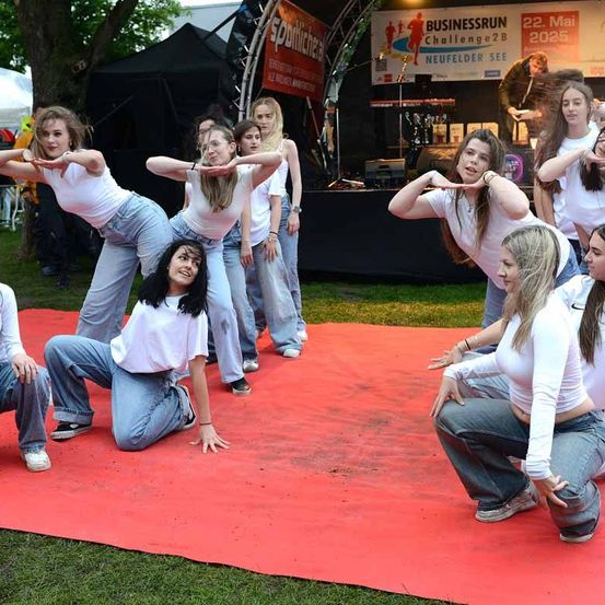 Eine Gruppe von Frauen in weißen Shirts und Jeans führt einen koordinierten Tanz auf einem roten Teppich auf, mit einer Bühne und Musikequipment im Hintergrund.