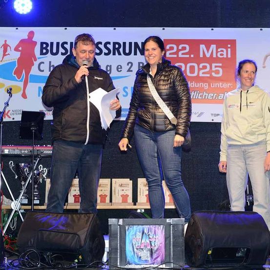 Ein Mann spricht in ein Mikrofon auf der Bühne, neben ihm stehen zwei Frauen. Hinter ihnen ist ein Banner mit dem Text Business Run Challenge.