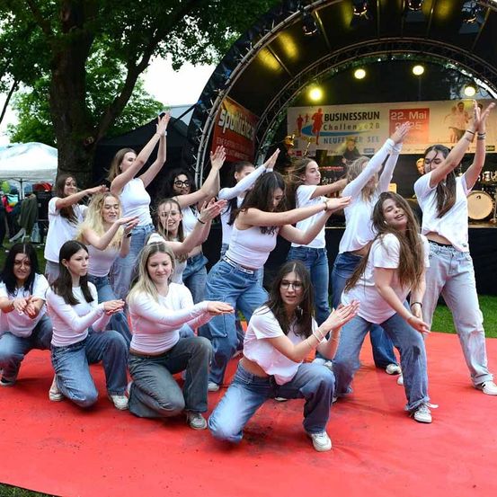 Eine Gruppe von Frauen in weißen Shirts und Jeans führt eine Tanzroutine auf einem roten Teppich auf. Hinter ihnen ist eine Bühne mit einem Banner zu sehen, auf dem 'Businessrun Challenge 2022' steht.