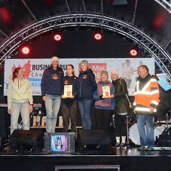 Eine Gruppe von Menschen steht auf der Bühne und hält Urkunden in den Händen. Sie sind in Winterkleidung gekleidet. Auf der Bühne befinden sich Lautsprecher und Trommeln. Hinter ihnen ist ein Banner mit Bildern von Menschen, die laufen.