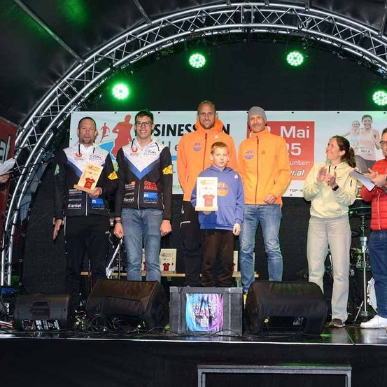 Mehrere Personen stehen auf einer Bühne und halten Trophäen und Urkunden. Hinter ihnen ist ein Banner mit einer Läuferfigur und Text zu sehen. Zwei Lautsprecher stehen auf der Bühne, und Lichter sind am Bogen darüber befestigt.