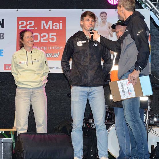 Ein Mann und eine Frau stehen auf der Bühne, der Mann wird von einem anderen Mann mit einem Mikrofon interviewt. Hinter ihnen steht ein Drumset und eine Banner ist angebracht.