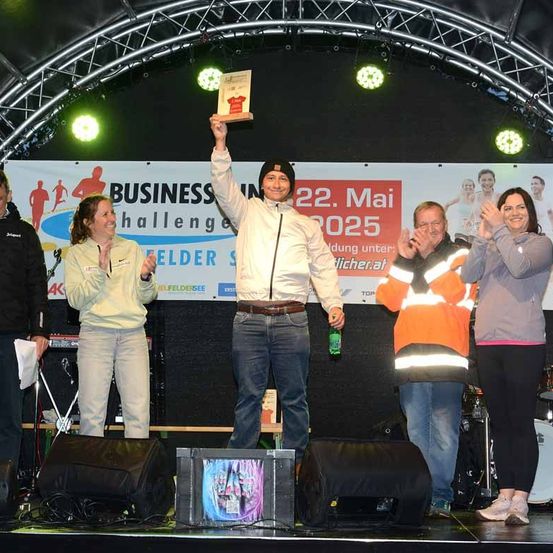 Ein Mann mit einer Plakette steht auf der Bühne mit drei Frauen und zwei Männern. Sie alle lächeln. Hinter ihnen ist ein Banner mit dem Text 'BUSINESS CHALLENGE'.