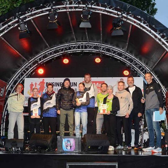 Sieger eines Rennens stehen auf einer Bühne und halten Pokale. Hinter ihnen zeigt ein Banner ein Logo und Text. Scheinwerfer sind oben und Lautsprecher unten.