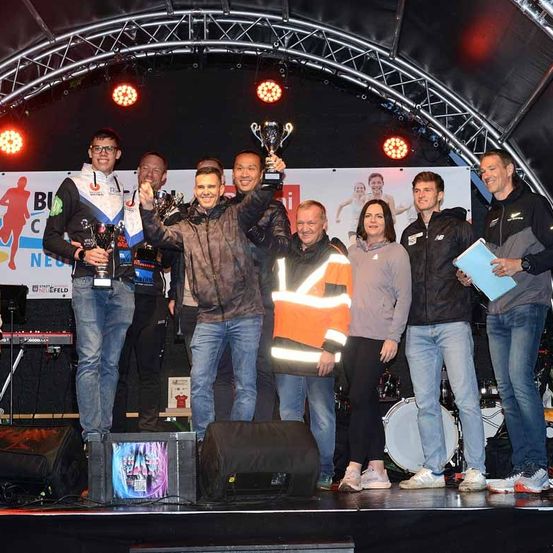 Mehrere Personen stehen auf einer Bühne, halten Pokale und lächeln für die Kamera. Dahinter ist ein Banner mit Logo und Text zu sehen. Musikinstrumente und Lautsprecher stehen auf der Bühne.