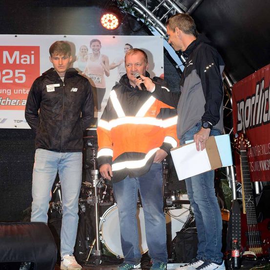 Drei Männer stehen auf einer Bühne. Ein Mann mit einem Mikrofon hält eine Mappe, während ein anderer Mann einen Drumstick hält. Ein Banner ist hinter ihnen angebracht.