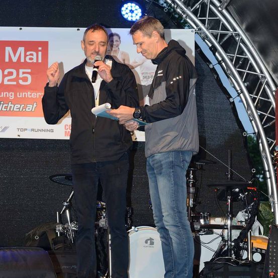 Zwei Männer auf einer Bühne, einer hält ein Mikrofon und spricht, während der andere ein Papier in der Hand hält und ihn ansieht. Dahinter steht ein Schlagzeug und ein Banner als Hintergrund.