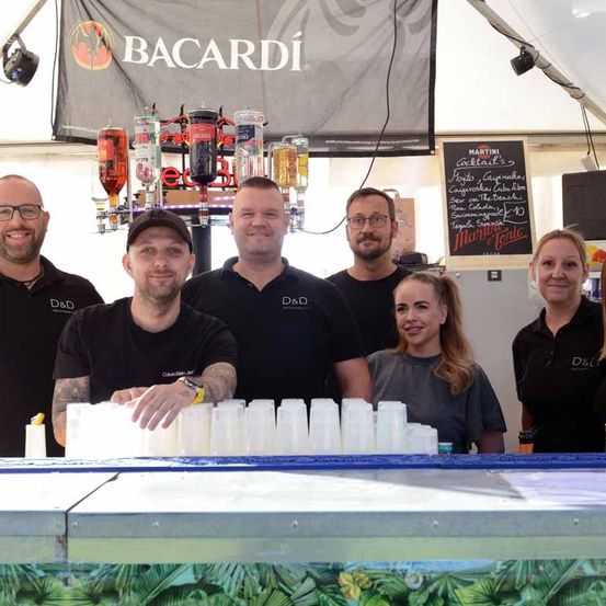 Eine Gruppe von Mitarbeitern steht hinter einer Bar mit einem Bacardi-Banner. Sie tragen schwarze Polo-Shirts. Vor ihnen befinden sich gestapelte Becher und eine Speisekarte.