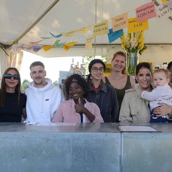 Eine Gruppe von Menschen steht unter einem Zelt und lächelt für ein Foto. Dahinter hängen bunte Banner mit Getränkepreisen. Eine Frau hält ein Baby.