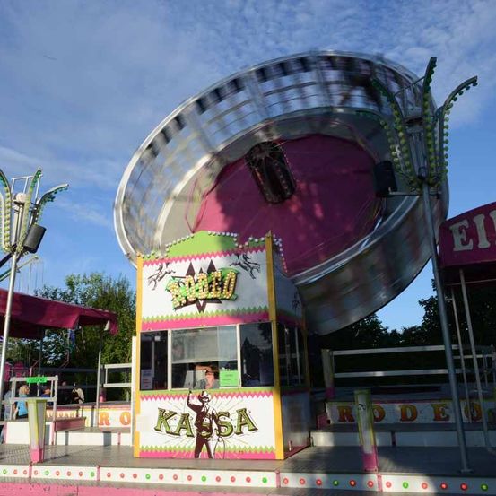 Ein Outdoor-Jahrmarktattraktion namens 'Kalsa' mit einer drehenden Fahrt im Hintergrund. Der Stand hat ein farbenfrohes Design mit einer Silhouette einer Person. Der Himmel ist blau mit einigen Wolken.