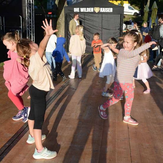 Eine Gruppe von Kindern tanzt auf einer hölzernen Bühne, mit einem Baldachin im Hintergrund, auf dem der Text 'Die Wilden Kaiser' steht. Einige tragen Turnschuhe, andere sind barfuß. Ein Mann steht in der Nähe des Baldachins.