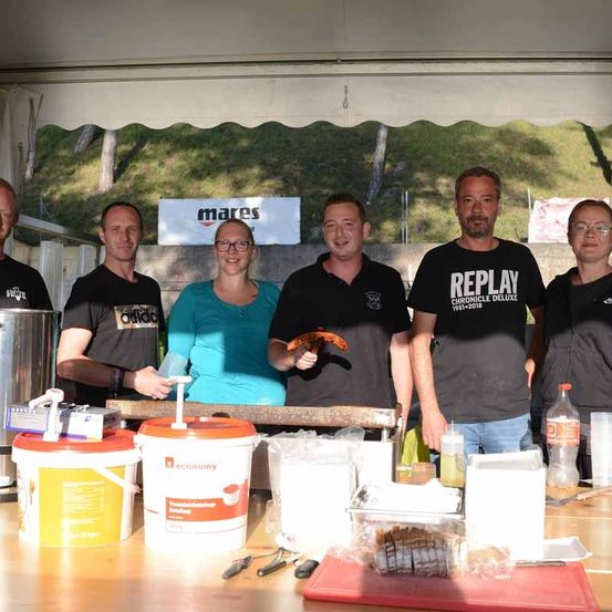 Fünf Personen posieren vor einem Tisch mit verschiedenen Behältern, Eimern und anderen Gegenständen. Hinter ihnen befindet sich ein grasbewachsenes Feld mit einem Schild, auf dem Mares steht.