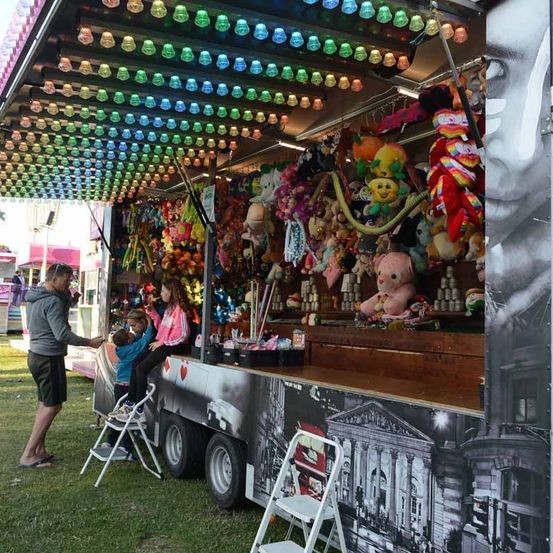Ein Karnevalsspielstand mit bunten Deckenlichtern und verschiedenen Kuscheltieren zur Schau gestellt. Ein Mann hilft zwei Kindern auf Leitern, um Preise zu erreichen.