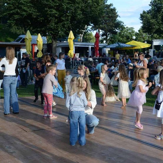 Eine Gruppe von Kindern tanzt auf einer hölzernen Tanzfläche unter gelben Sonnenschirmen. Einige Erwachsene beobachten sie. Bäume, Straßenlaternen und mehrere Personen, die auf Bänken sitzen, sind im Hintergrund zu sehen.