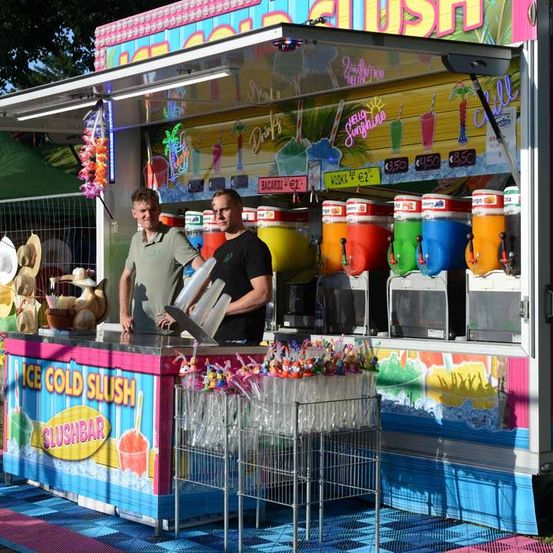 Zwei Männer stehen hinter einem Eis-Kälte-Slush-Bar, mit bunten Slushies und einem Regal mit Strohhalmen. Der Bar ist hell dekoriert mit verschiedenen Fruchtgetränken und einer großen Menütafel.