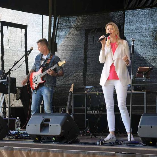 Eine Frau singt auf der Bühne, während ein Mann Gitarre spielt. Hinter ihnen steht ein Mikrofon und Musikequipment aufgebaut.